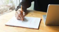 Hand marking a date on a desk calendar next to a laptop.